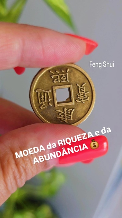 Moeda Feng Shui dourada com inscrições chinesas segurada entre dedos com unhas vermelhas