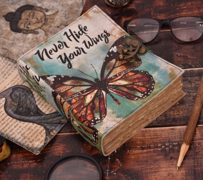Caixa de livro decorativa com borboleta e texto 'Never Hide Your Wings' numa mesa de madeira com acessórios