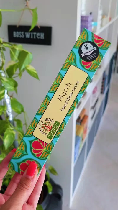 Caixa colorida de incenso Natural Masala Aurah com padrão floral, segurada à frente, ambiente interior com planta e placa BOSS WITCH ao fundo