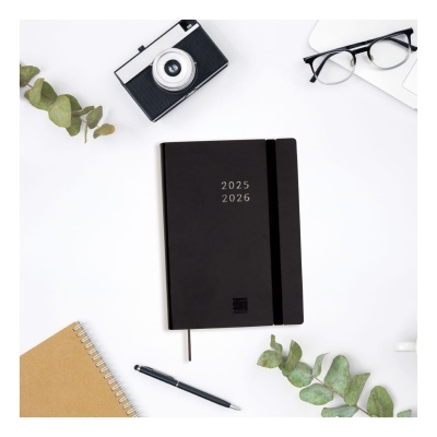 Agenda preta 2025 2026 com elástico, câmara preta e branca, óculos, caderno castanho, caneta e folhas verdes