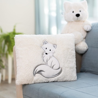 Almofada felpuda bege com bordado de raposa branca sobre sofá azul e urso de peluche no fundo