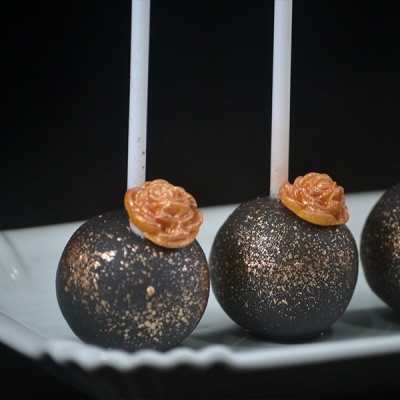 Three black and gold-speckled cake pops with orange flower decoration on a white plate