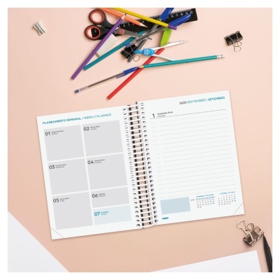 Agenda semanal espiralada aberta com artigos de papelaria sobre mesa cor de rosa