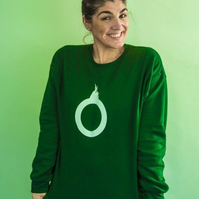 Mulher a usar camisola verde escura com círculo branco e chama, texto 'SÓS AMA', fundo verde claro.