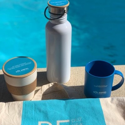 Garrafa metálica branca com tampa de cortiça, caneca azul e copo reutilizável com faixa de cortiça com marca SOS ANIMAL junto a toalha azul perto de piscina