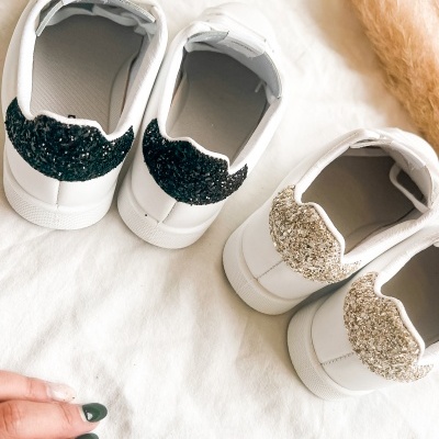 Dois pares de sapatilhas brancas com glitter na parte de trás, vistas de trás, sobre tecido branco.