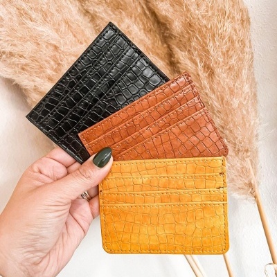 Carteiras porta-cartões em preto, castanho e amarelo com textura de pele de crocodilo seguradas numa mão.
