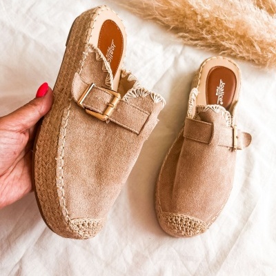 Sapatos mule bege com fivela dourada e sola em fibra natural num fundo branco com plumas