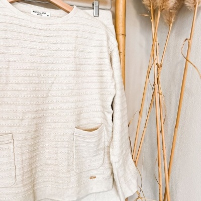Conjunto de camisola creme com padrão e bolsos, pendurado em cabide de madeira num cabideiro de bambu