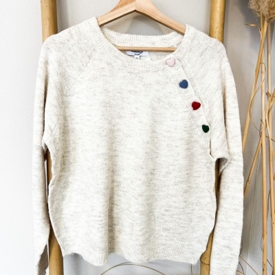 Camisola creme com botões de coração coloridos no ombro, pendurada em cabide de madeira numa estrutura de bambu.