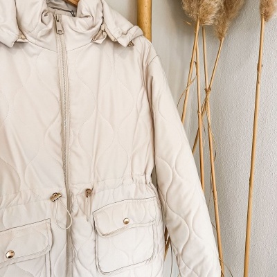 Casaco bege acolchoado com bolsos e fecho de correr pendurado em cabide de madeira.