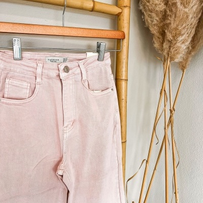 Calças de ganga cor-de-rosa penduradas em cabide de madeira, parede branca e flores secas ao lado