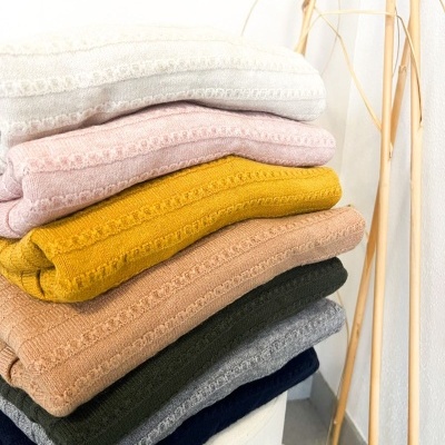 Mantas ou toalhas de várias cores empilhadas com textura e padrão, em ambiente minimalista com chão de azulejos e varas decorativas.