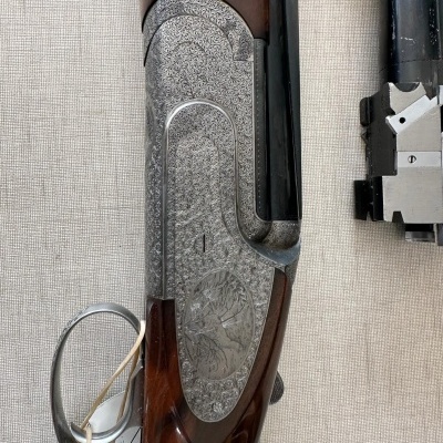 Espingarda de caça com madeira e metal gravado exibida sobre fundo claro com etiqueta