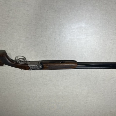Espingarda de canos duplos com coronha em madeira escura e canos pretos contra parede clara
