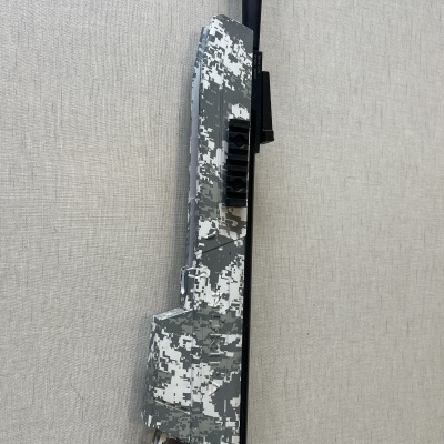 Rifle de ar comprimido camuflado digital cinzento, branco e preto contra parede cinzenta