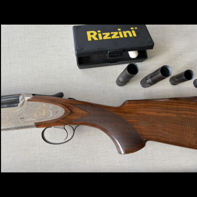 Espingarda com coronha em madeira polida, caixa preta com o texto Rizzini e quatro tubetes metálicos.