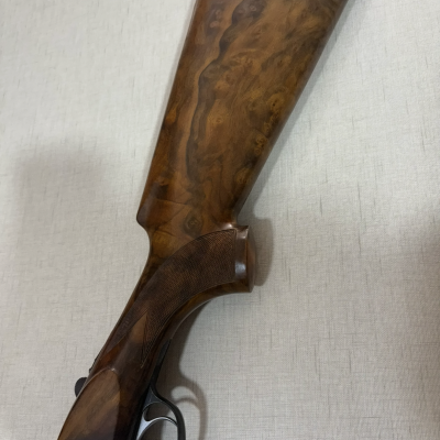 Coronha de espingarda em madeira castanha com detalhes em metal num fundo cinza.