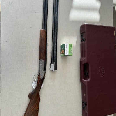 Espingarda desmontada com madeira castanha e canos pretos, caixa de munições verde e estojo vermelho sobre superfície clara