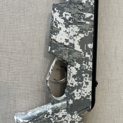 Arma com padrão camuflado digital em tons de cinza e branco num fundo cinza claro