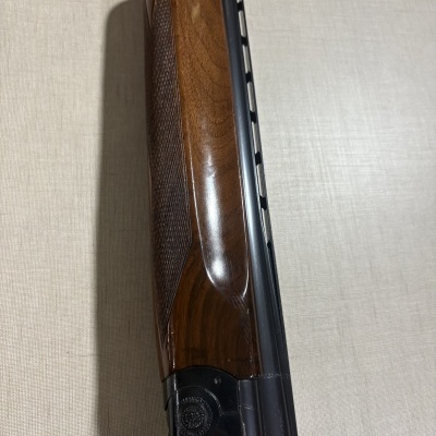 Parte de espingarda com coronha em madeira e cano metálico preto numa superfície bege.