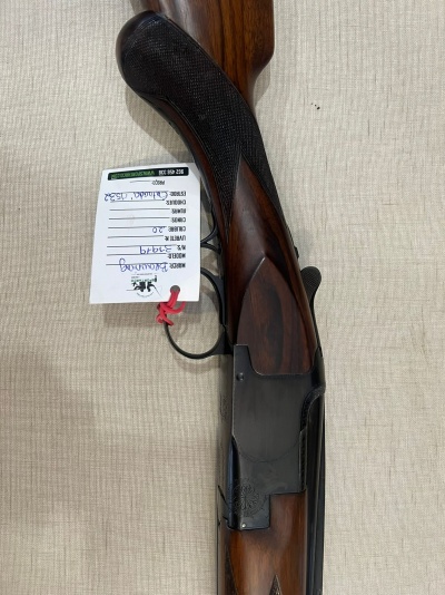 Parte inferior de espingarda de cano duplo com cabo em madeira castanha e metal negro, etiqueta branca pendurada.