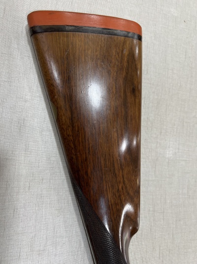 Cabo de espingarda em madeira com borracha vermelha e detalhe preto sobre tecido bege.