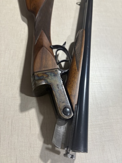 Espingarda de caça de canos duplos em metal negro e madeira com textura quadriculada, mostrada aberta.