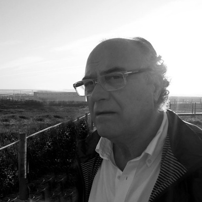 Homem de meia-idade com óculos e casaco ao ar livre junto a uma cerca em área aberta.