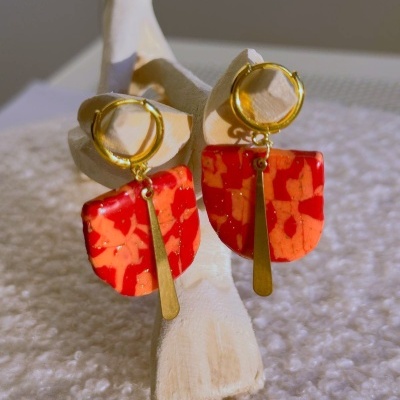 Brincos pendentes dourados com base vermelha e laranja e pendente metálico em suporte claro.
