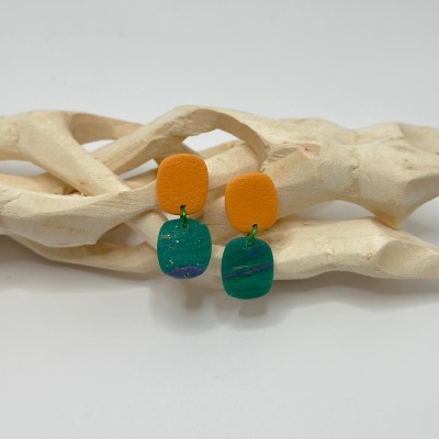 Brincos laranja e verde com detalhes em roxo e dourado sobre estrutura de madeira clara