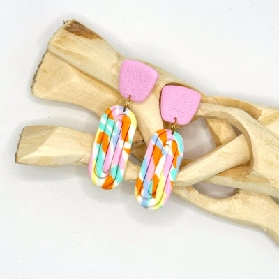 Brincos coloridos com base rosa e parte inferior oval colorida sobre madeira clara.