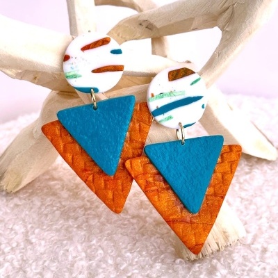 Brincos pendentes circulares brancos com padrões coloridos e triângulos azuis e laranja, exibidos em suporte de madeira clara