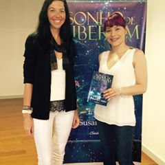 Duas mulheres em frente a banner promocional, uma segura um livro