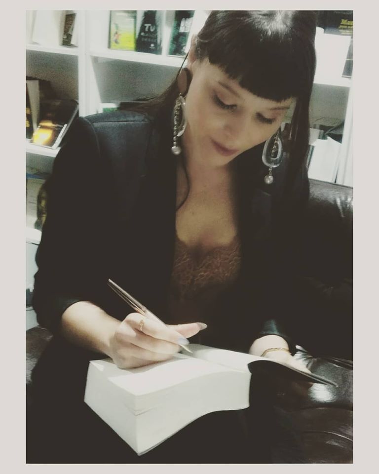 Feira do Livro - Porto Mulher a assinar livro grosso com casaco preto e brincos grandes sentada num sofá escuro