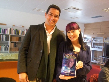 Duas pessoas em ambiente interior com estantes de livros, mulher a segurar livro 'SONG OF LIBERATION'