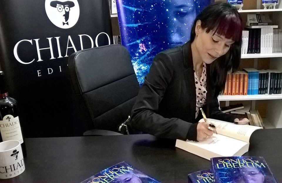 Mulher a assinar livro numa mesa preta com livros, garrafa, copo, cadeira, estante de livros e painel Chiado Editora