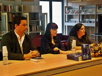 Três pessoas sentadas numa mesa comprida com livros e garrafas de água numa sala com estantes de livros