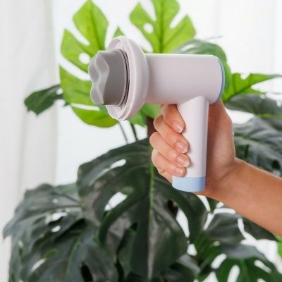 Dispositivo portátil branco e azul com ponta cinzenta em formato de pétalas, segurado por uma mão com folhas verdes ao fundo