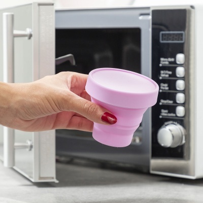 Mão segurando copo de silicone rosa em frente a micro-ondas