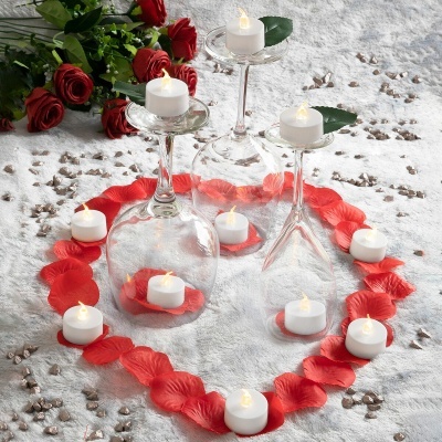 Copos de vidro com velas e pétalas formando um coração ao lado de um ramo de rosas vermelhas