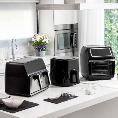 Três fritadeiras de ar pretas numa bancada de cozinha branca moderna com flores num vaso ao fundo