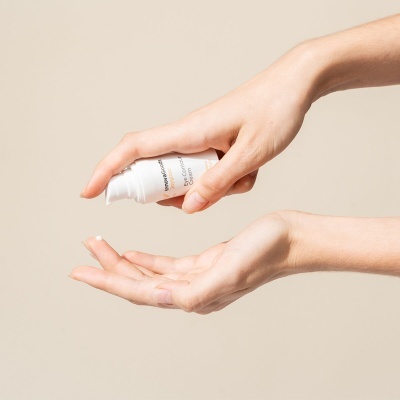 Mãos aplicando creme de embalagem branca sobre fundo bege.