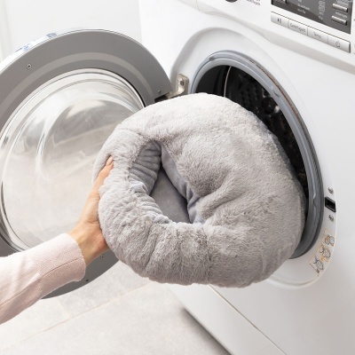 Máquina de lavar branca com almofada cinzenta peluda sendo colocada dentro.