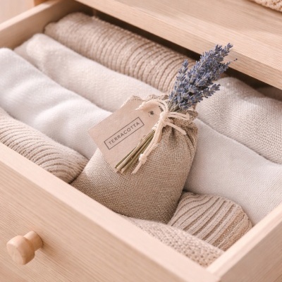 Gaveta de madeira com roupas de malha creme e bege e saco de tecido com ramo de flores secas roxas e etiqueta TERRACOTTA