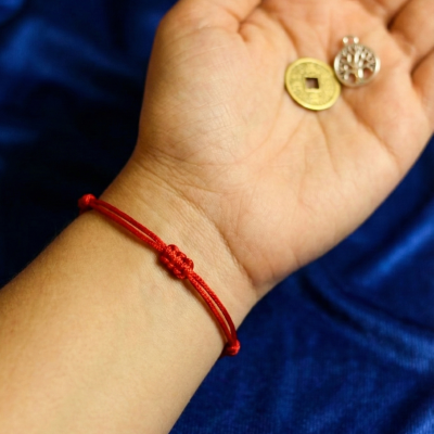 Pulseira vermelha de cordão com nó central na mão segurando moeda dourada e pendente prateado sobre tecido azul.