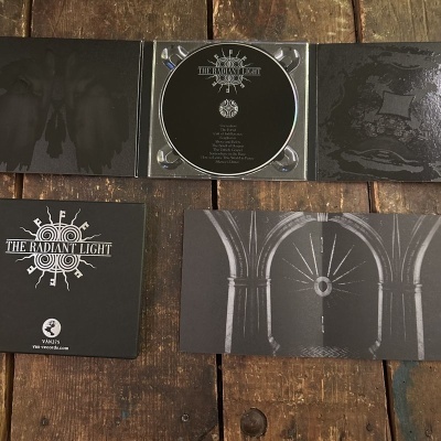 CD preto e embalagem com texto THE RADIANT LIGHT sobre mesa de madeira