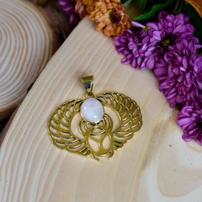 Pendente dourado com pedra branca sobre madeira ao lado de flores roxas e laranjas