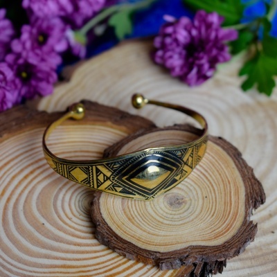 Pulseira dourada com padrão tribal sobre pedaços de madeira e flores roxas