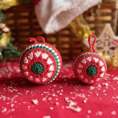 Dois ornamentos de croché vermelho, verde e branco com padrão circular, sobre tecido vermelho com neve artificial.